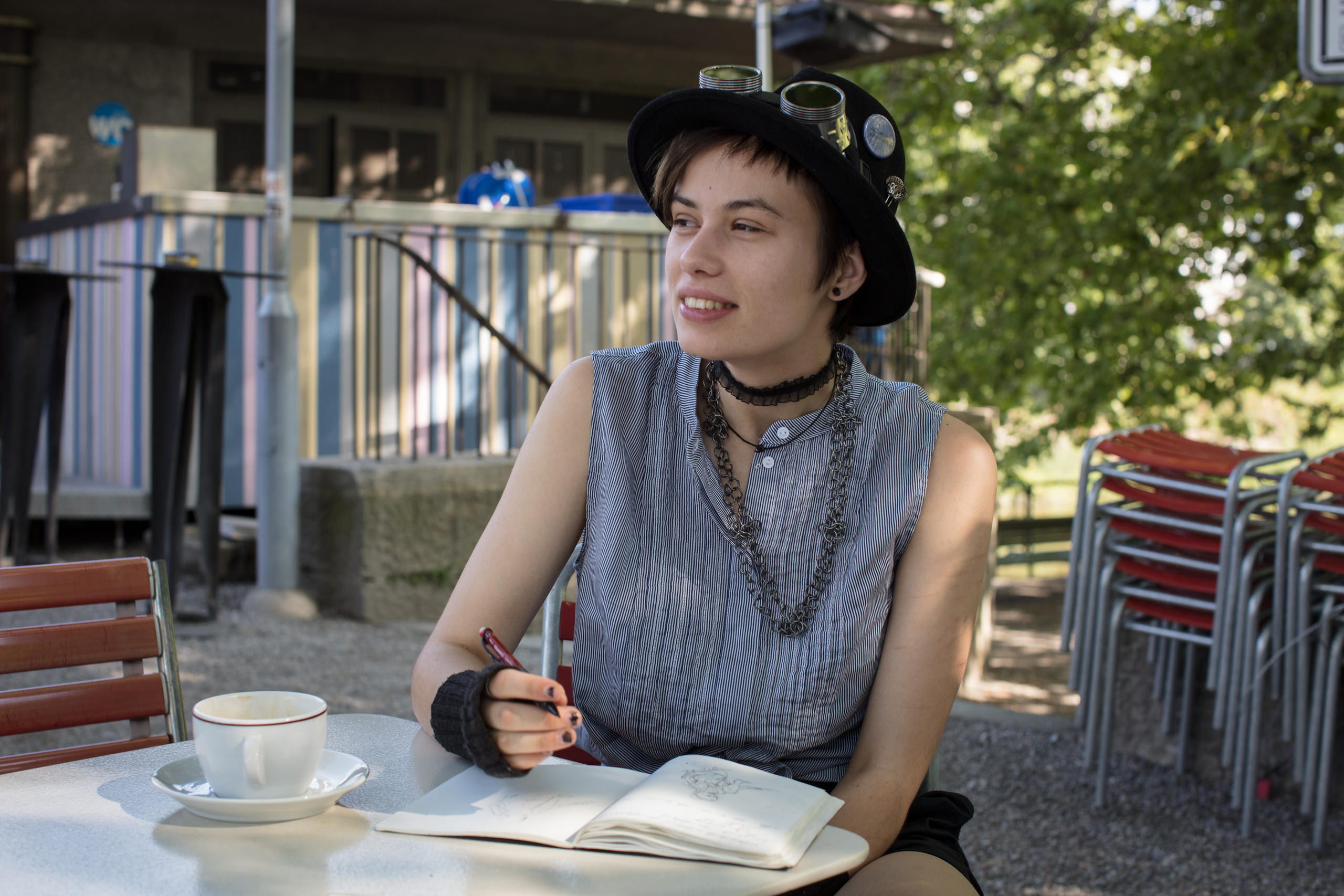 Eine Frau mit Hut sitzt in einem Garten an einem Tisch, vor ihr eine Kaffeetasse und ein Heft.