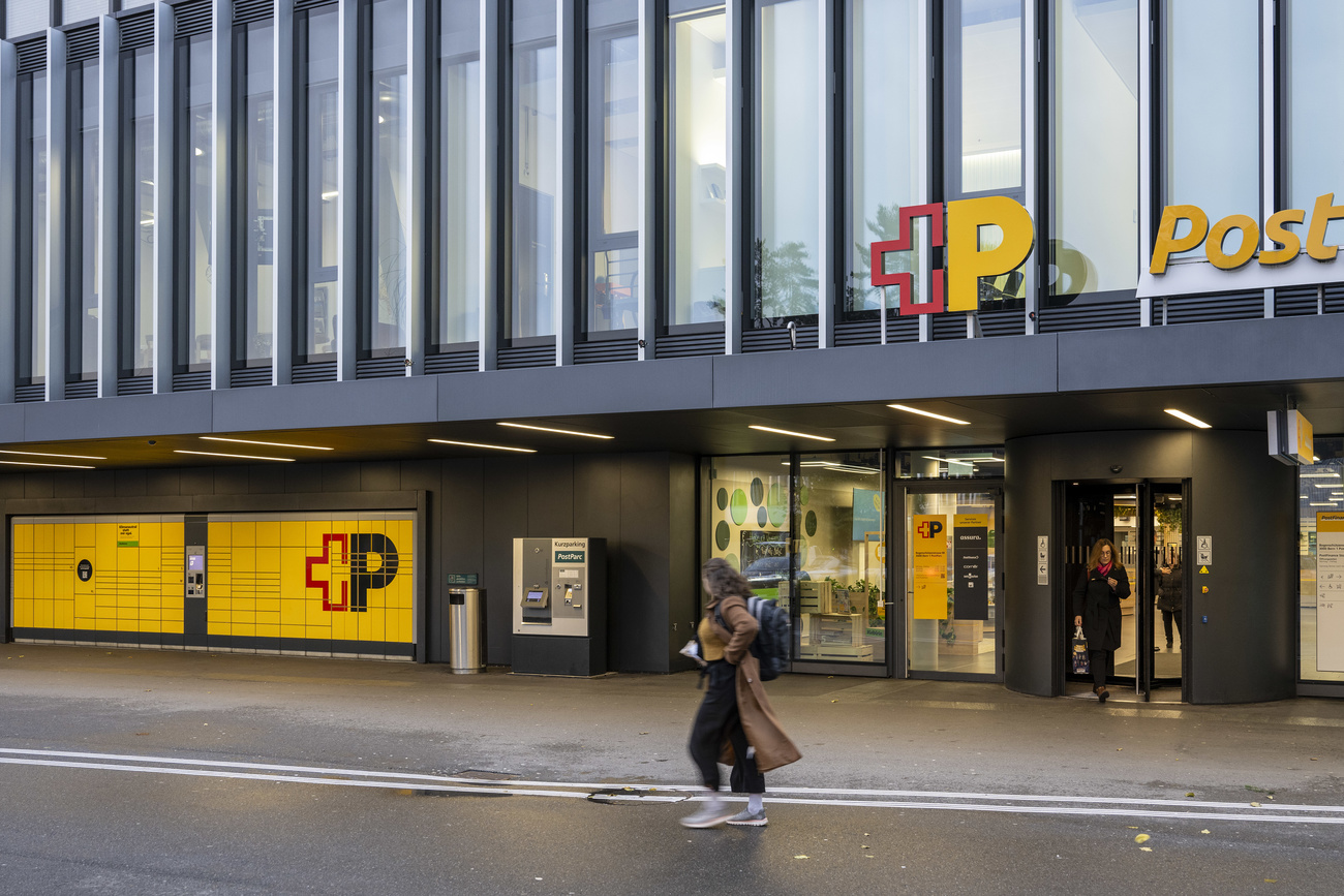 A woman in a brown coat walks in the road. Behind her is a large Swiss Post branch, with the ‘+P’ logo in red (+) and yellow (P) above the door and a wall of bright yellow Post lockers