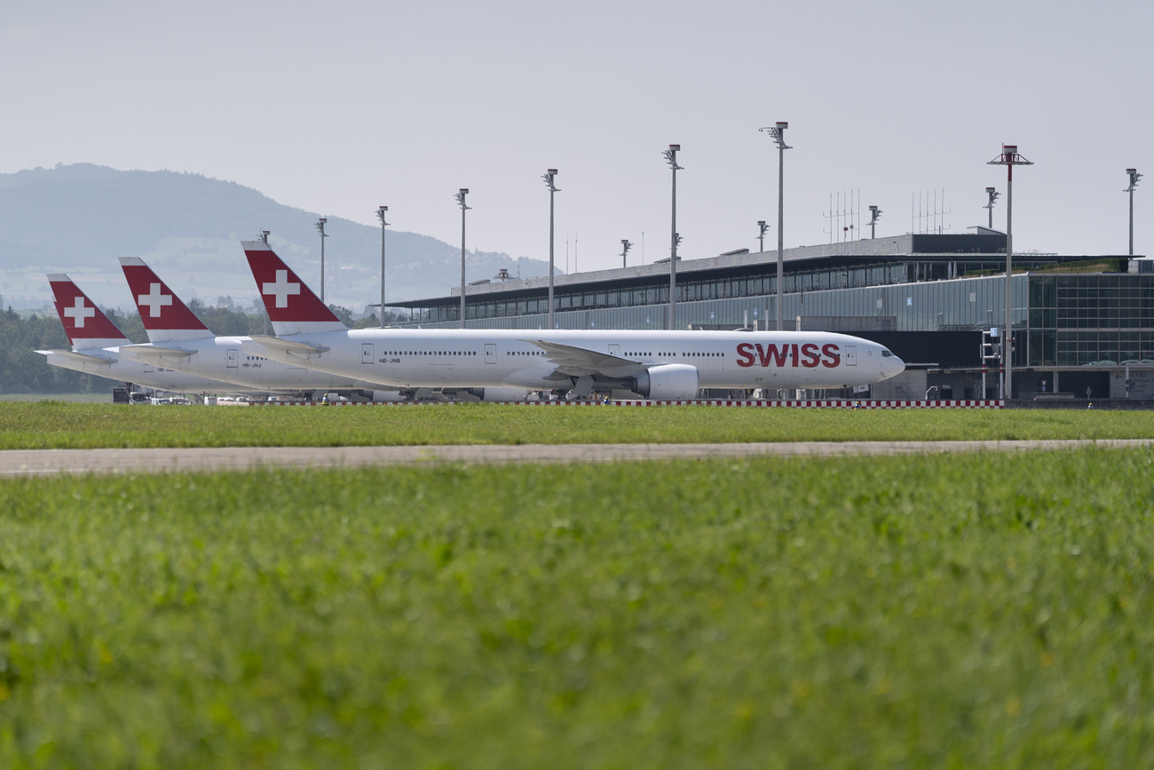 Three SWISS planes are lined up at Zurich airport in front of the airport building, the red Swiss flag ‘plus’ on their tails visible one behind the other, as well as ‘SWISS’ in big letters on the foremost aircraft. Mountains can be seen in the distance and in the foreground of the image is a grassy area.