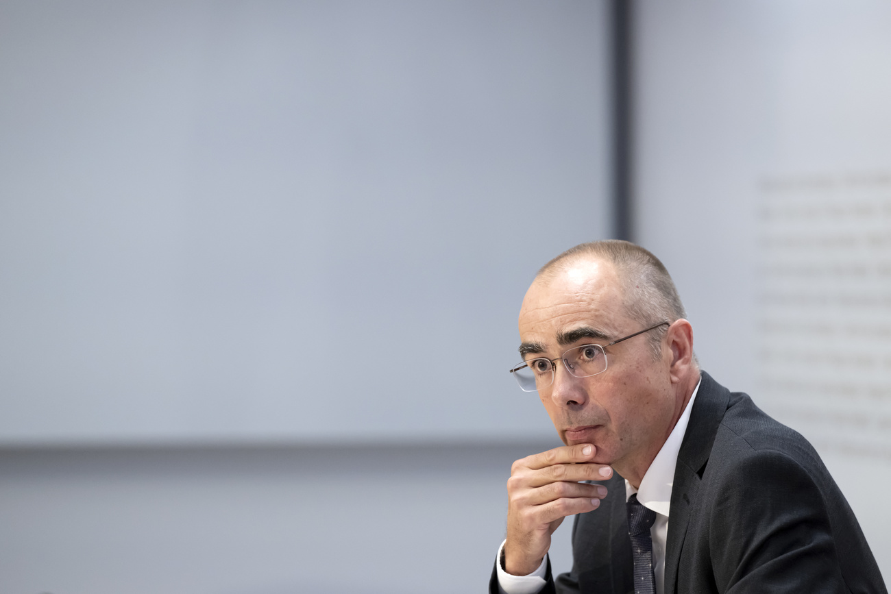 New head of customs, Pascal Lüthi, sits rubbing his chin in a white room. He is wearing a dark suit jacket, a black tie, a white shirt and glasses, and has short, grey hair