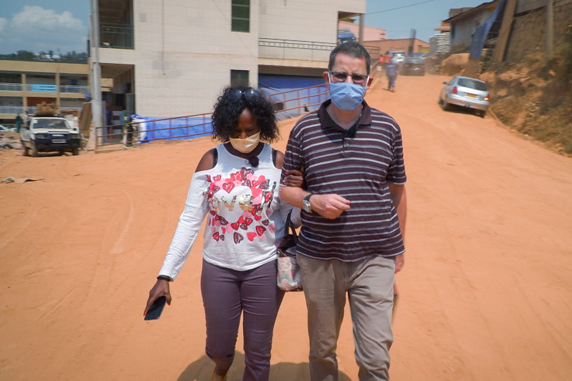 Homme blanc et femme noire se tenant pas le bras dans une rue en Afrique avec un masque covid sur le visage