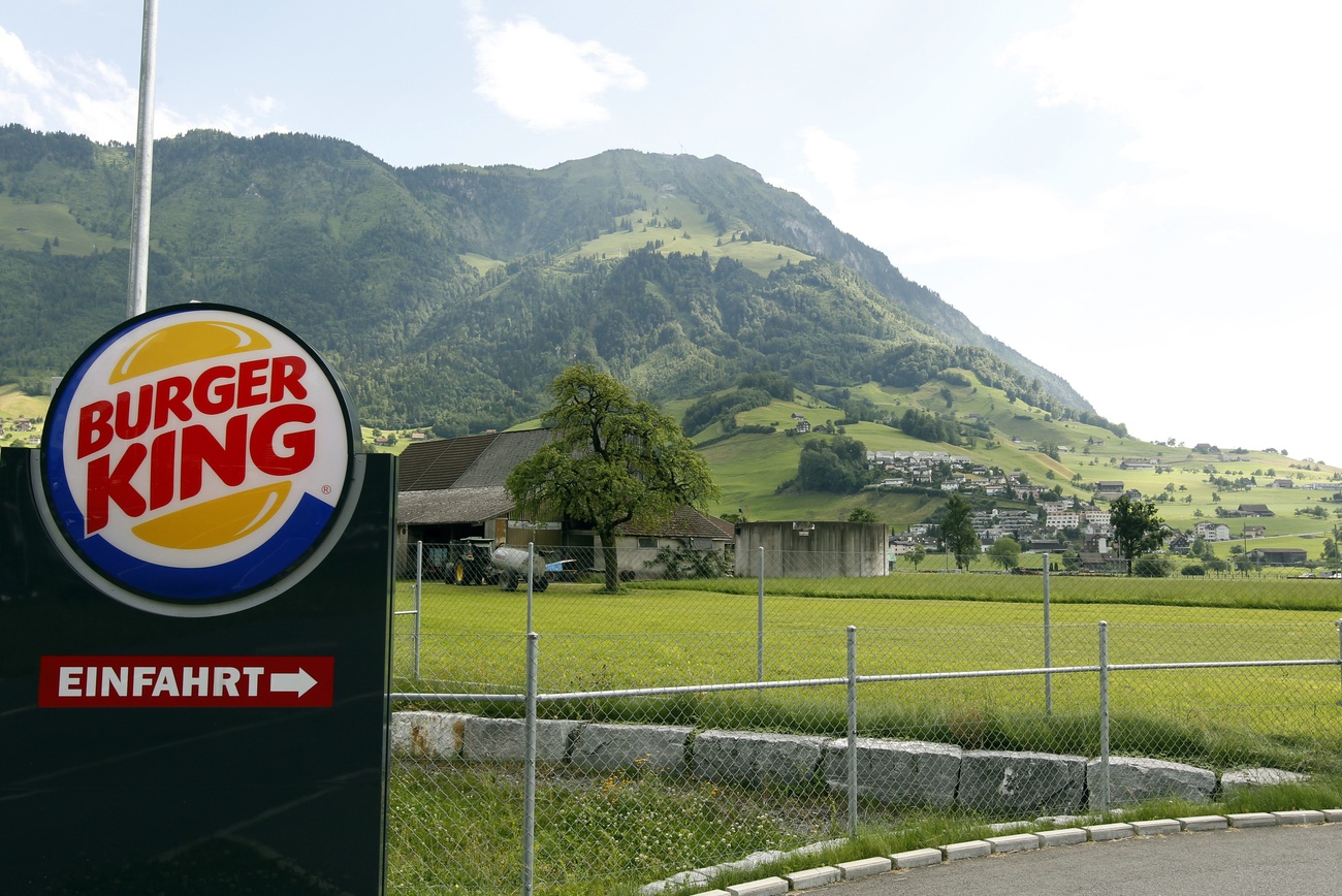 A black Burger King drive-thru entrance sign with the Burger King logo on the top: a blue circle with white in the middle, and two yellow half-circle ‘buns’ above and below the words ‘Burger King’ in big red font. The sign is in front of a fence, behind which can be seen a green, mountainous Swiss landscape.