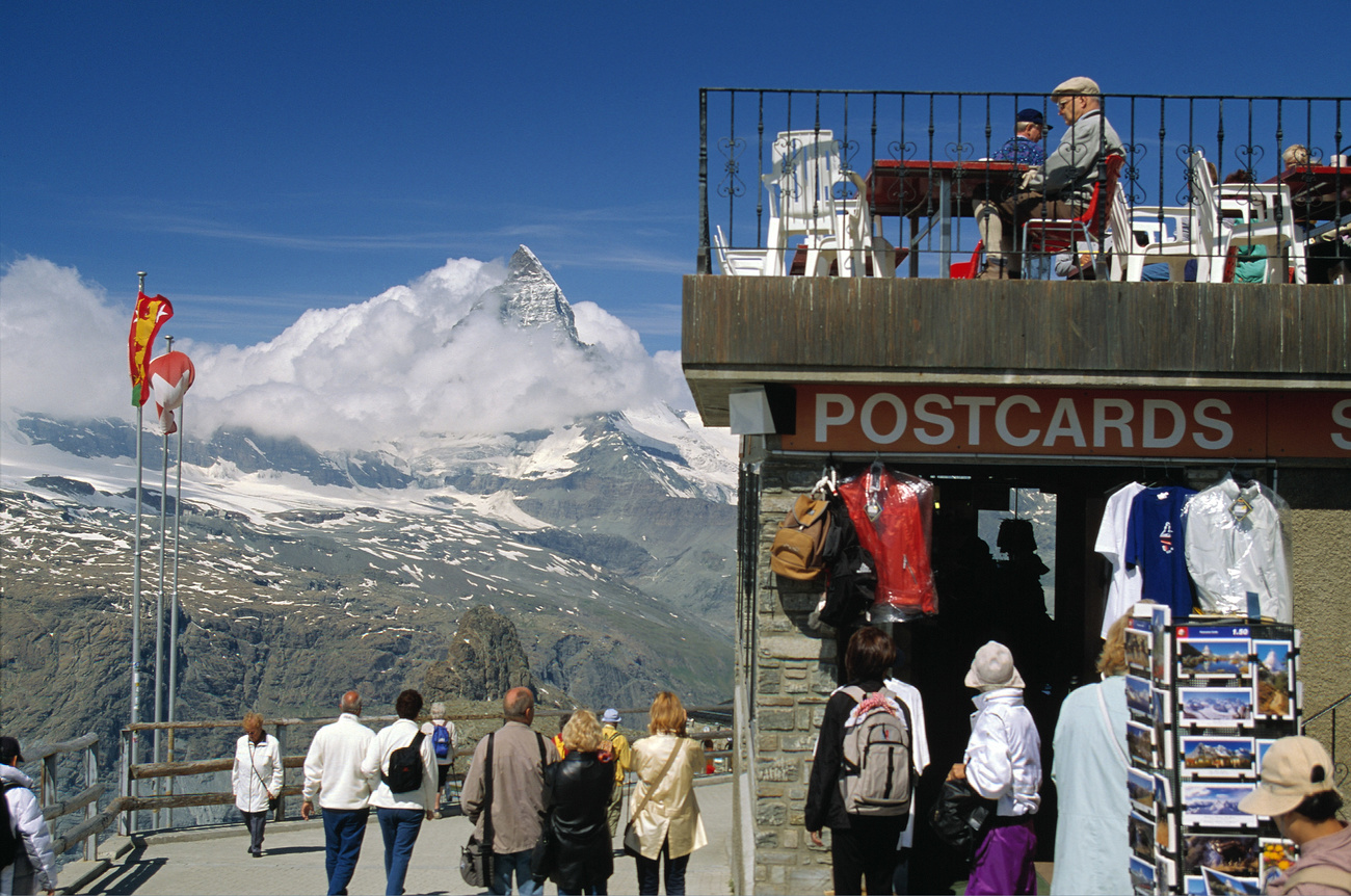 A number of tourists are seen walking near mountains and a sign on a building reading "postcards".