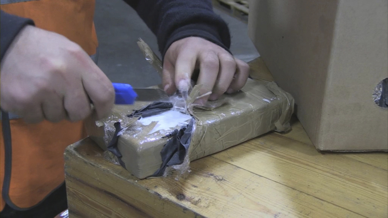 A pair of gloved hands are shown cutting into a package containing a large amount of cocaine.