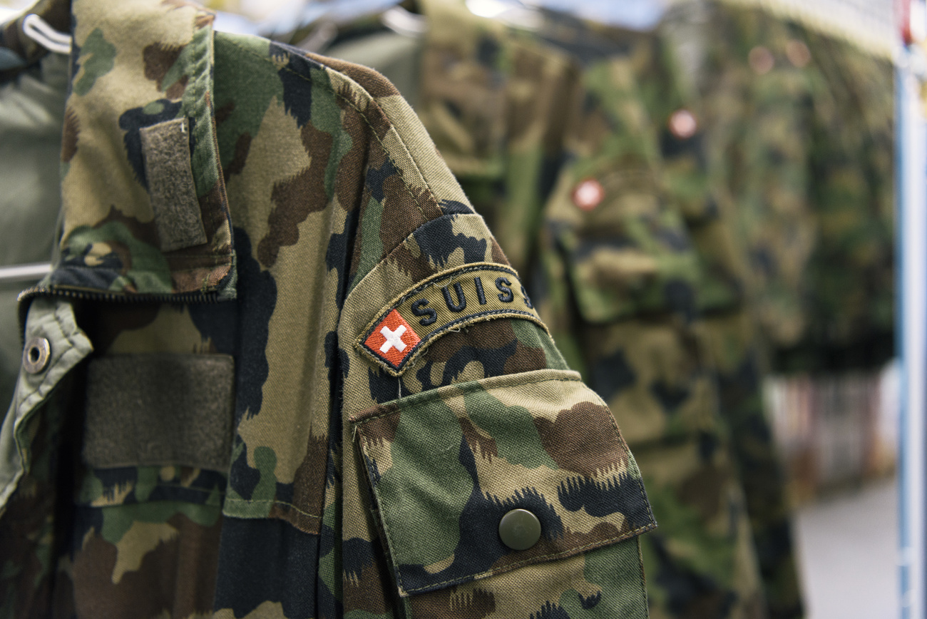 The top and sleeve of a camouflage Swiss armed forces shirt on a wire hanger. The red Swiss flag with a white plus is on the left shoulder with the word ‘Suisse’ above a breast pocket. Other uniforms can be seen hanging in the background of the image.