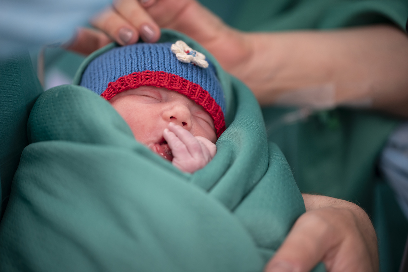 A baby yawns into its hand. It is wrapped in a teal blanket with a blue knitted hat on its head with a white flower and red trim. An adult’s arm can be seen cradling the child and another adult’s hand is touching its head.