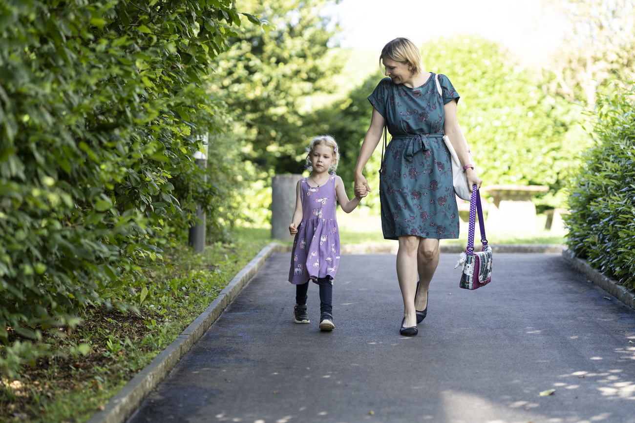 A mother holds hands with her daughter as they walk to the first day of kindergarten. The woman is dressed in a business outfit.