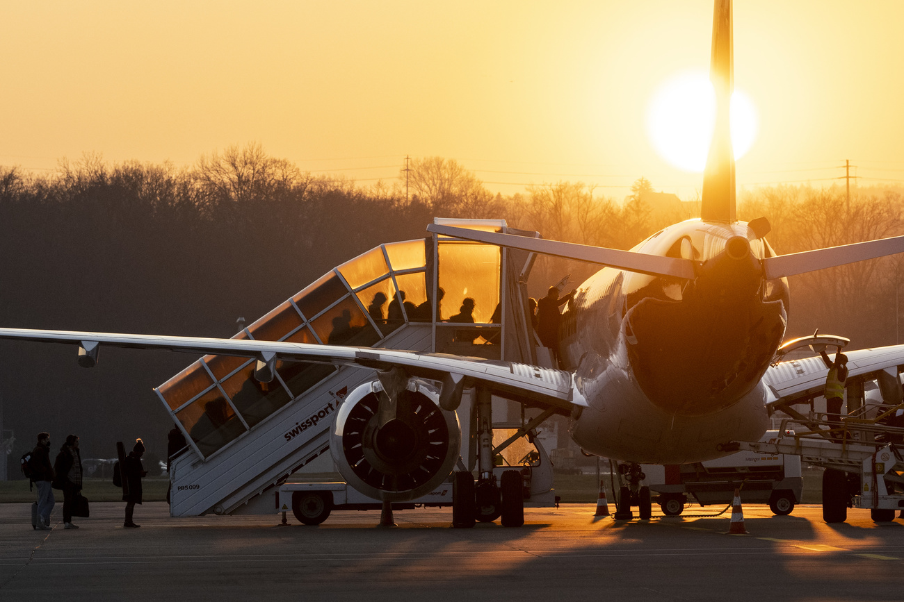 Passengers ascend a covered stairway into an aeroplane parked on a runway. The sun is setting, giving the scene a golden glow, with the sphere of the sun cut in two by the tail of the plane. The dark outline of trees can be seen in the background.
