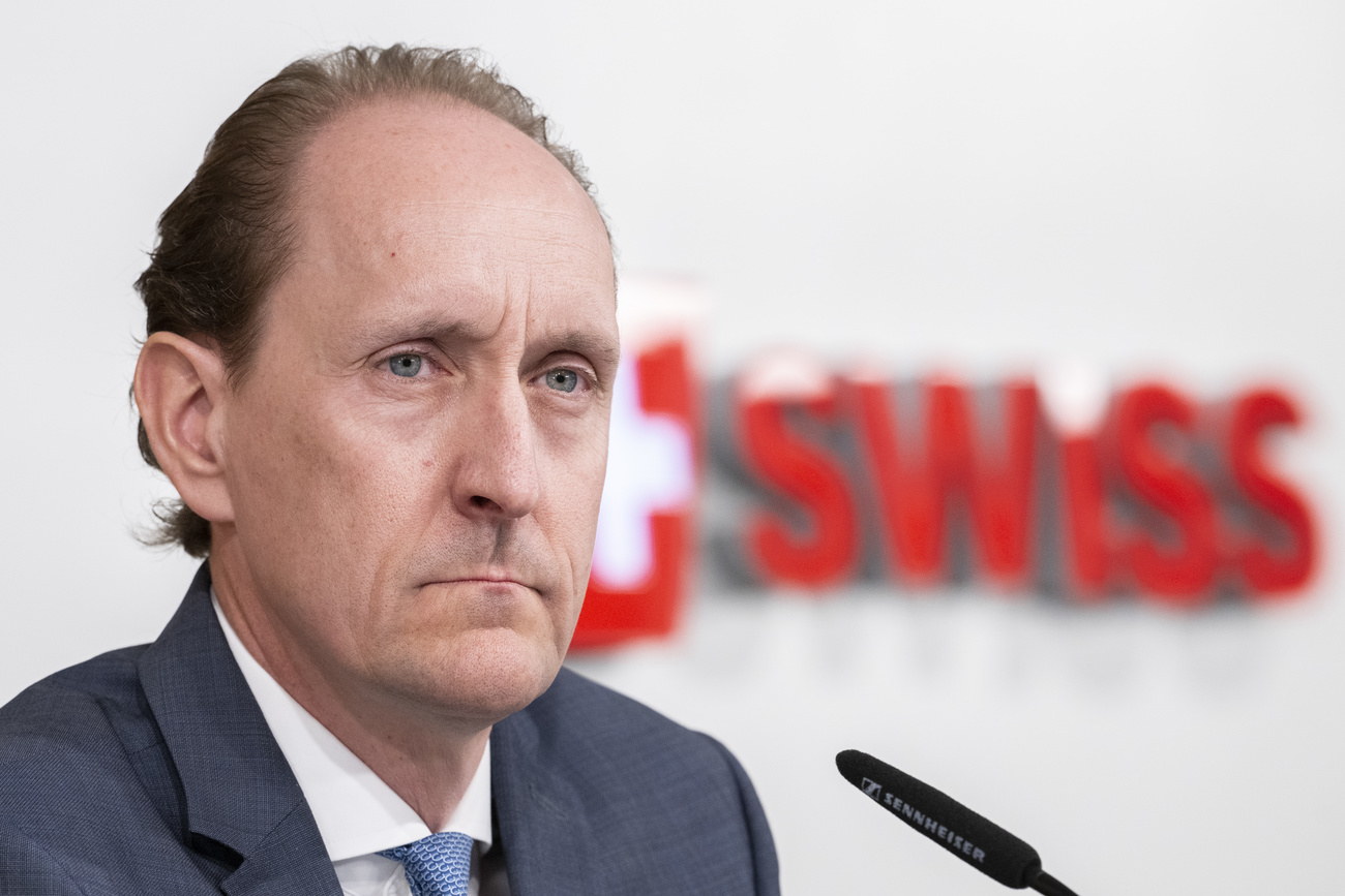 Swiss International Air Lines (SWISS) CEO Dieter Vranckx looks off to the right of the picture. He is frowning slightly and wearing a grey suit with a light blue tie and a white shirt. Behind him can be seen the blurred red SWISS logo.