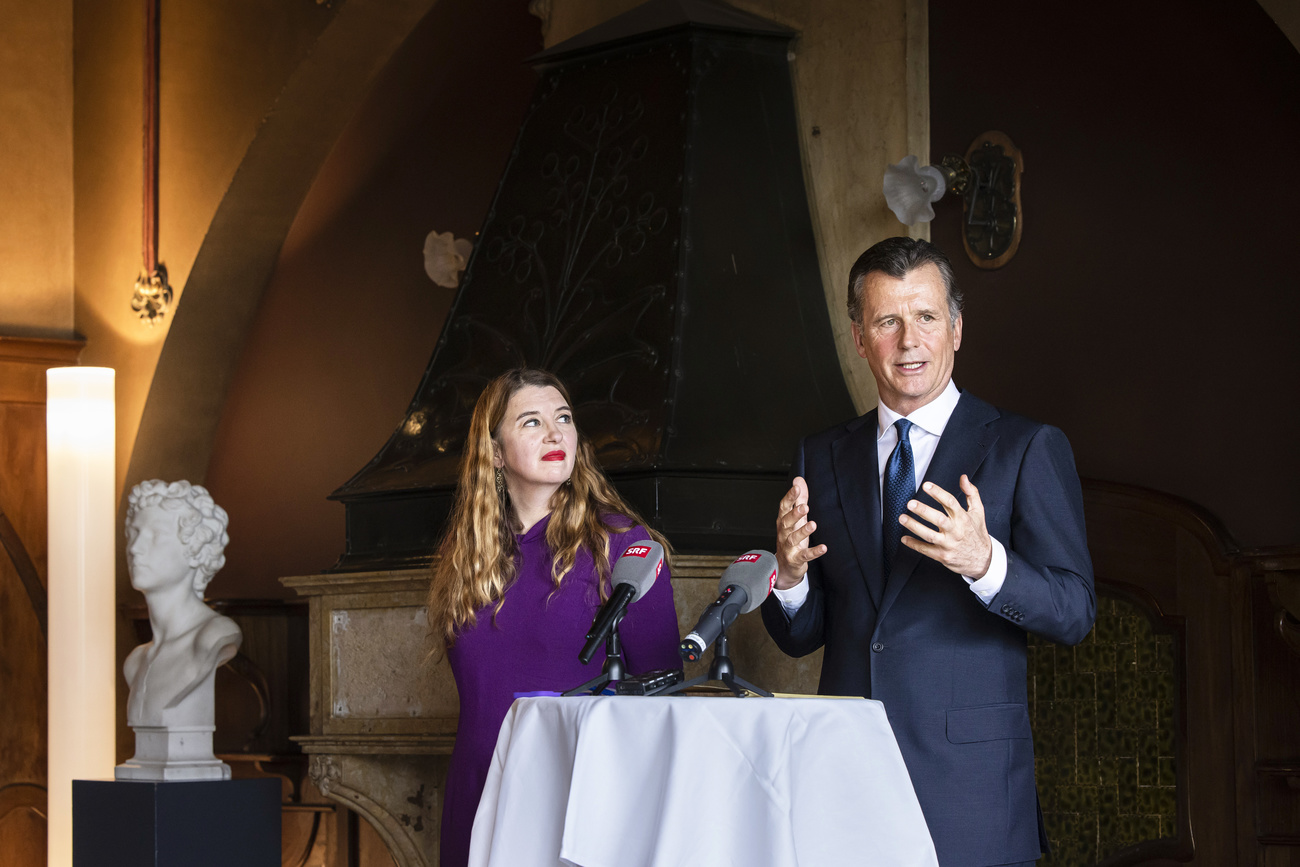 Philipp M. Hildebrand, President of Zurich Art Society, right, and Ann Demeester, Director of the Kunsthaus Zurich, left. Hildebrand wears a navy suit, a deep blue tie and a white shirt, and his hands are held out in front of him as he speaks. Demeester, who has long, curly, blonde hair and is wearing a purple dress, is watching him. They are stood behind a table with two SRF microphones on a white tablecloth. To their right is a white marble bust and a standing lamp. They are in an old room with stone walls.