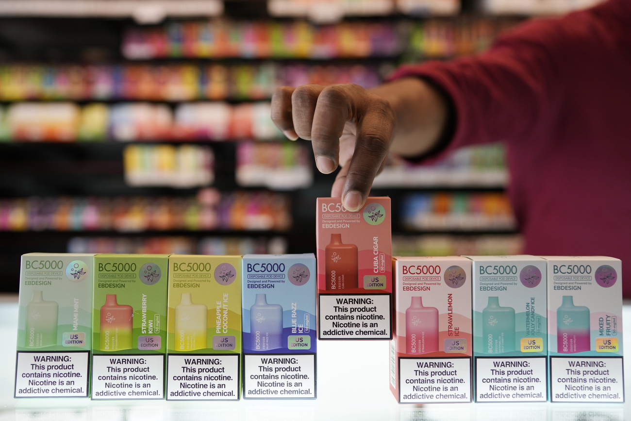 A line of disposable e-cigarette packets on an uplit shop counter. A person’s hand is picking up a red packet in the middle. There are several different coloured packets: red, green, yellow, pink, and all have nicotine warning labels on the front.