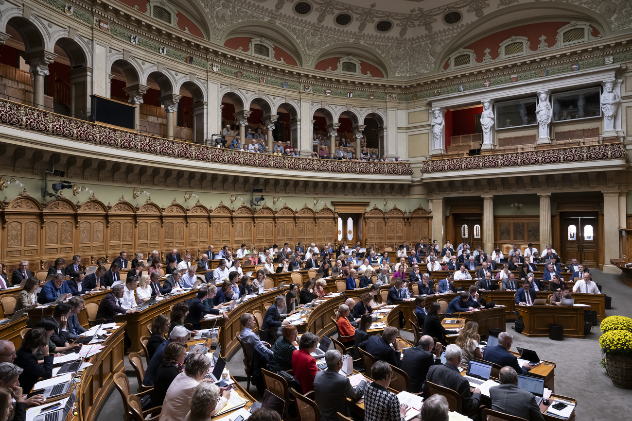 Bern's Nationalrat full of parliamentarians. Wooden desks curve in a semicircle around a central point to the left. The room is wood-panelled with ornate balconies and arches above.