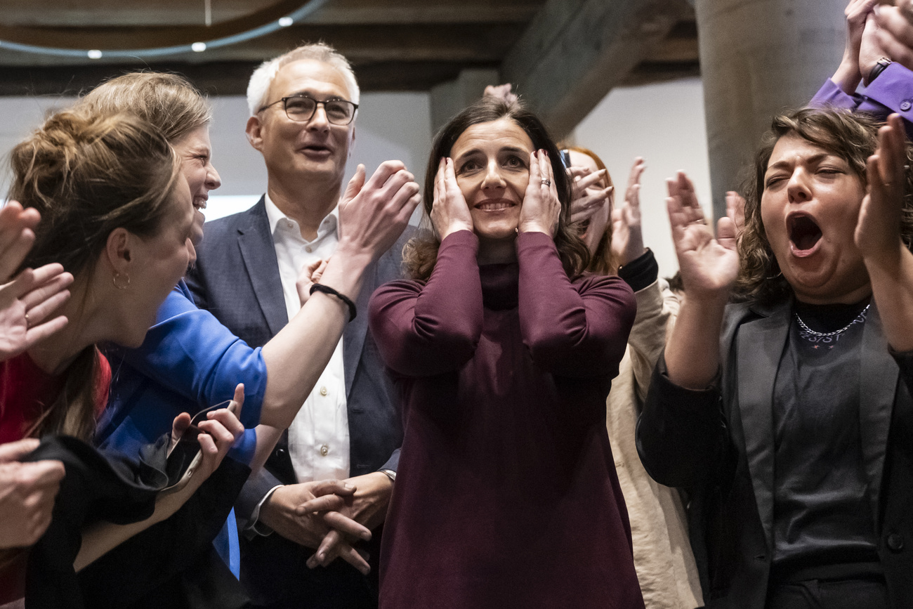 Social Democrats Senate candidate Flavia Wasserfallen reacts to the election announcement at the centre of the photo. She is smiling and both hands are on her face. Green candidate Bernhard Pulver is to her right. Those around them are smiling, cheering and clapping.
