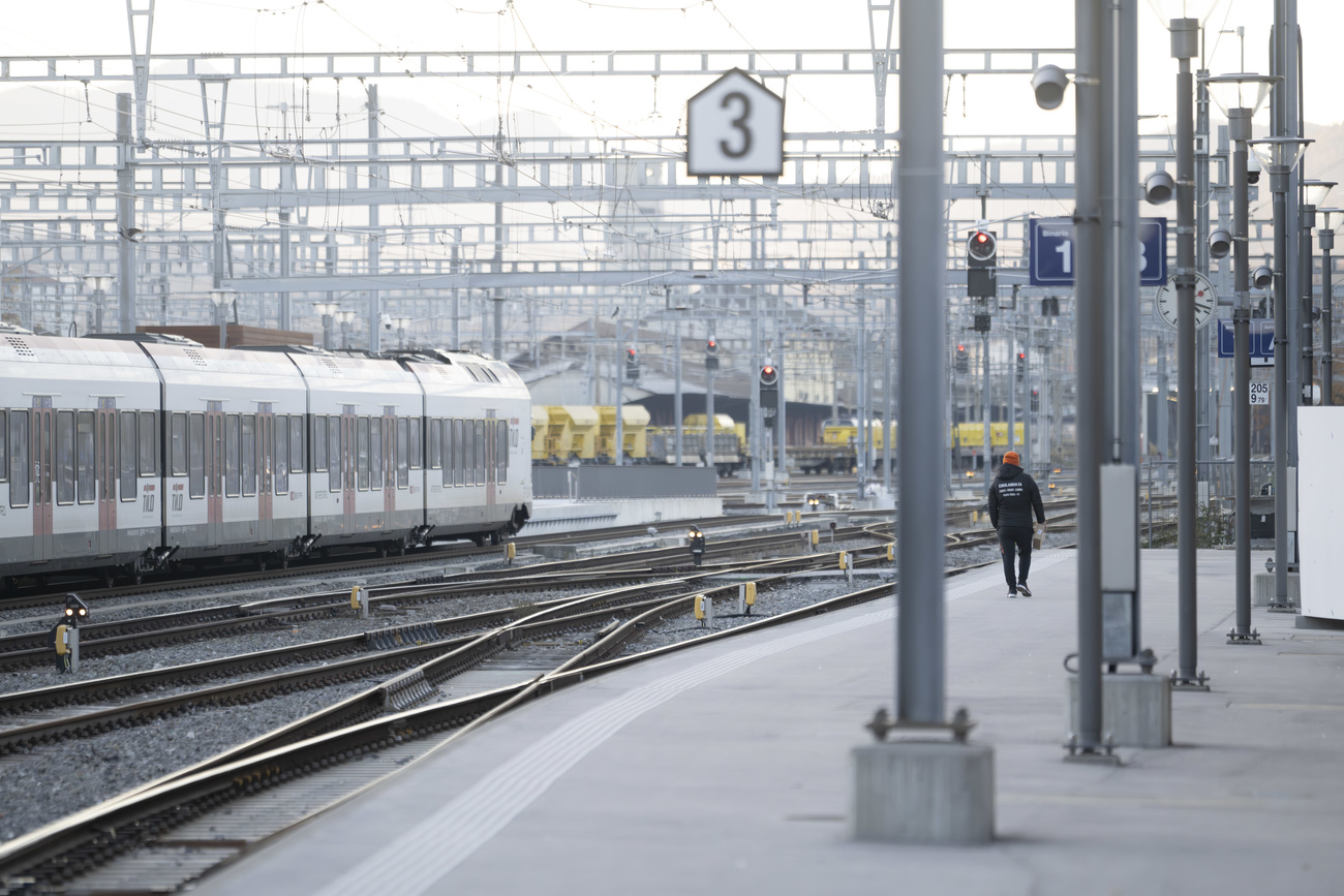 Platform 3 at Chiasso train station in Switzerland. The camera is looking down the platform, one man is walking along it but it is otherwise empty. A train can be seen pulling into a platform on the left. The train tracks and lines overhead are also visible on the left of the platform.