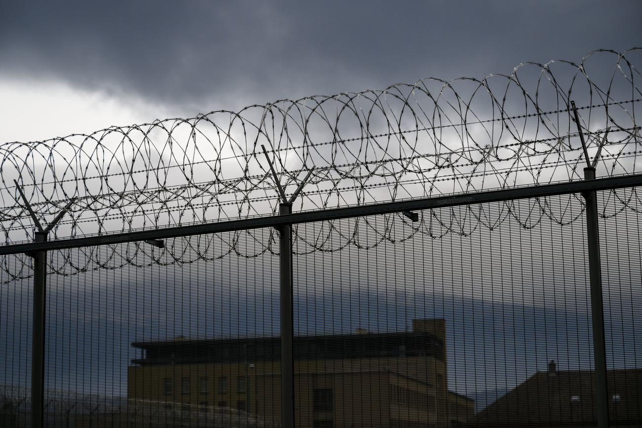 Rolls of barbed wire along the top of a fence. The shadows of buildings can be seen behind them, the sky is dark and cloudy.
