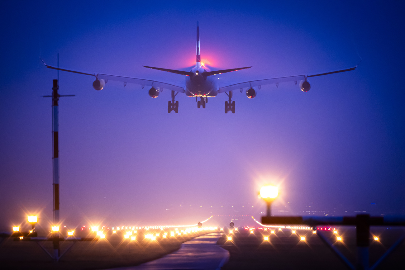 A plane comes in to land on the runway at Zurich airport. It is dark and the sky is a deep purple and the runway is dotted with yellow lamps.