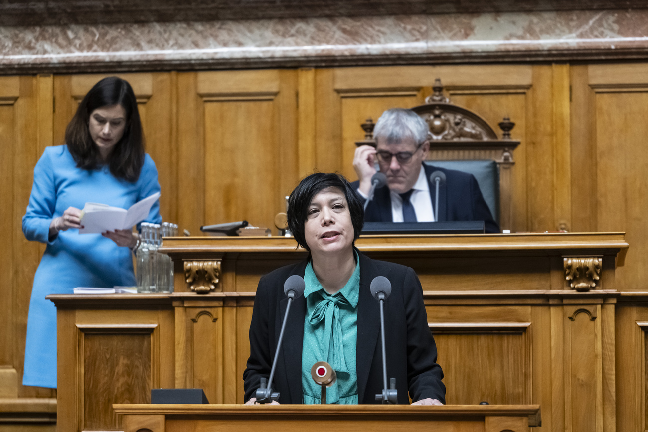 Min Li Marti speaking at a wooden podium in Switzerland’s wood-panelled House of Representatives. She has short hair and is wearing a black jacket with a jade green blouse with a bow at the neck, and is speaking into two microphones in front of her. Behind her in a blue dress is vice-president Maja Riniker to the left, and directly behind her seated in an ornate chair is president Eric Nussbaumer, who has short grey hair and is wearing a black suit and tie and a white shirt.