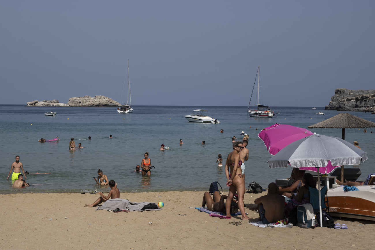 Tourists enjoy the beach and the sea in Lindos, on the island of Rhodes, Greece. There are people swimming in the sea and boats slightly further out, while on the beach people sit on towels under umbrellas.