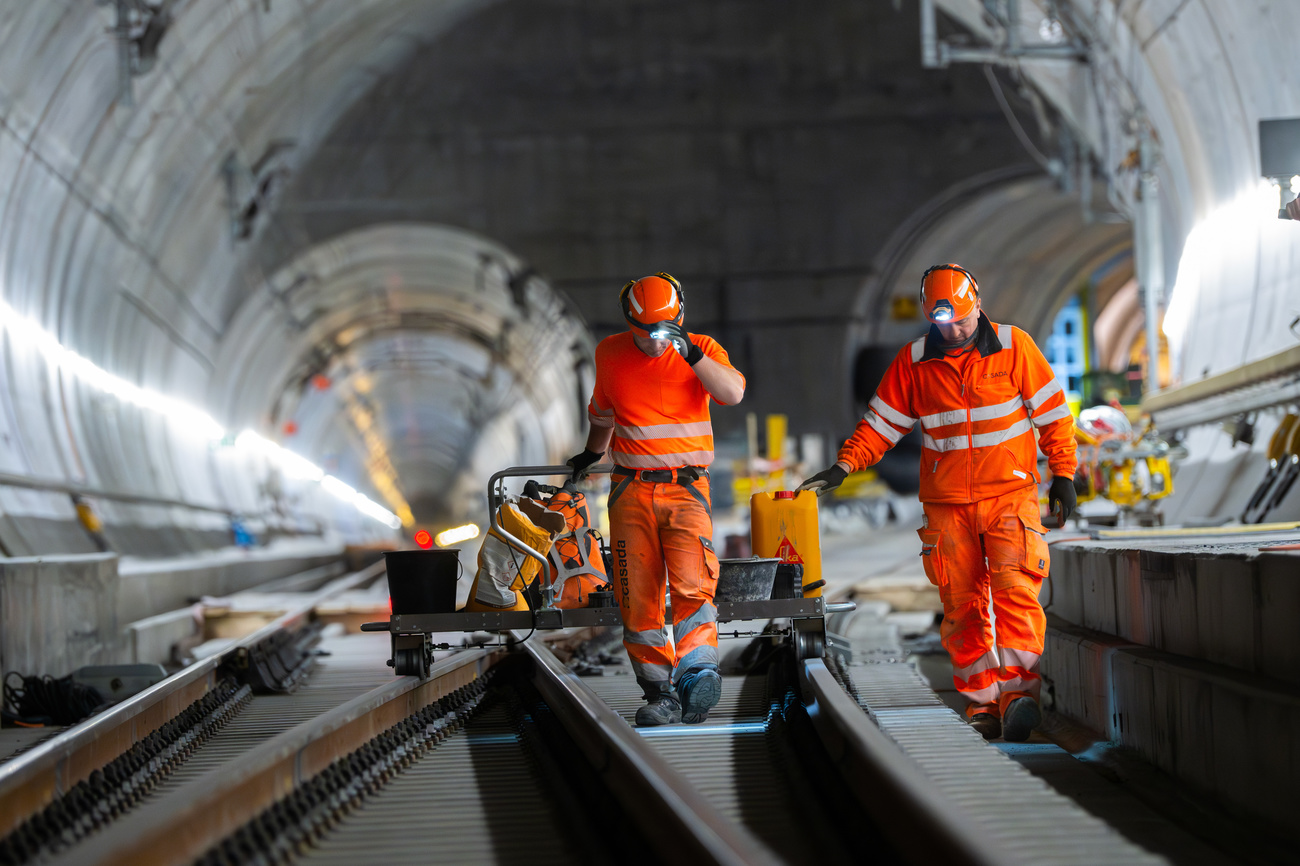 Maintenance workers wearing orange uniforms and hard hats with torches on the front walk towards the camera through the Gotthard tunnel. They are both looking at the ground. The tunnel looms huge above them and can be seen splitting into two tubes in the background.