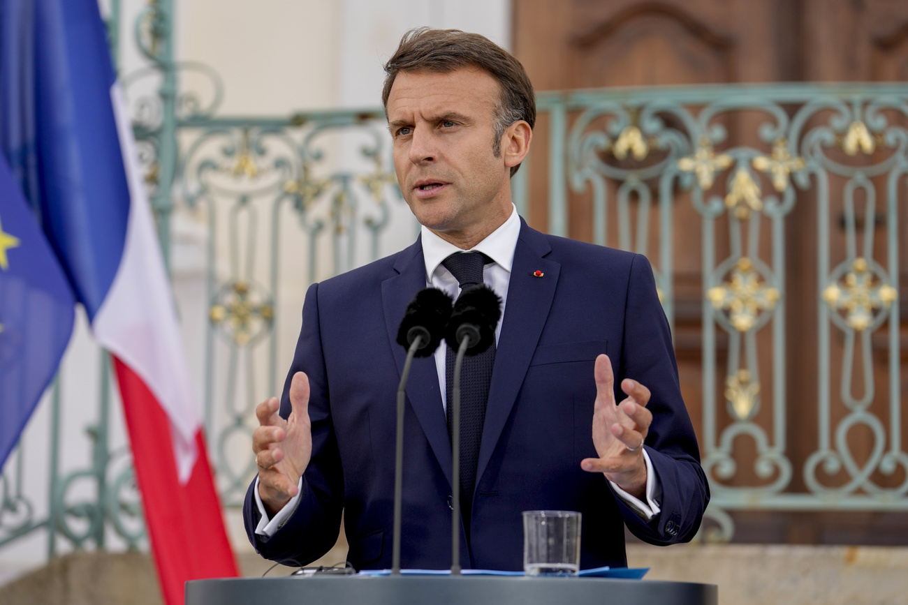 French President Emmanuel Macron speaks at a podium into a microphone. He is wearing a navy suit, a black tie and a white shirt. Behind him are a French flag and intricate railings.