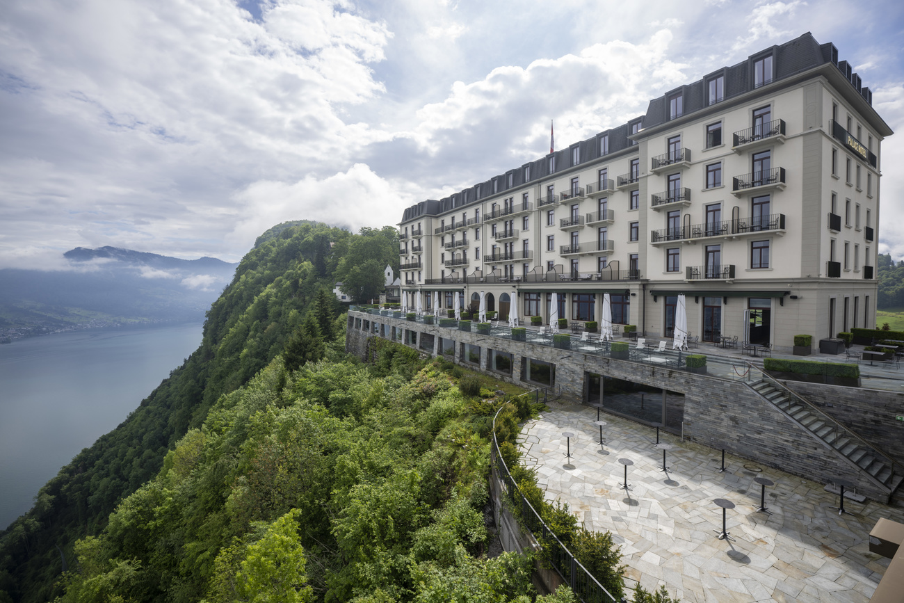 The Burgenstock resort, perched on the top of a tree-covered mountain. Lake Lucerne can be seen behind in the distance. The building is approximately six storeys, and white.