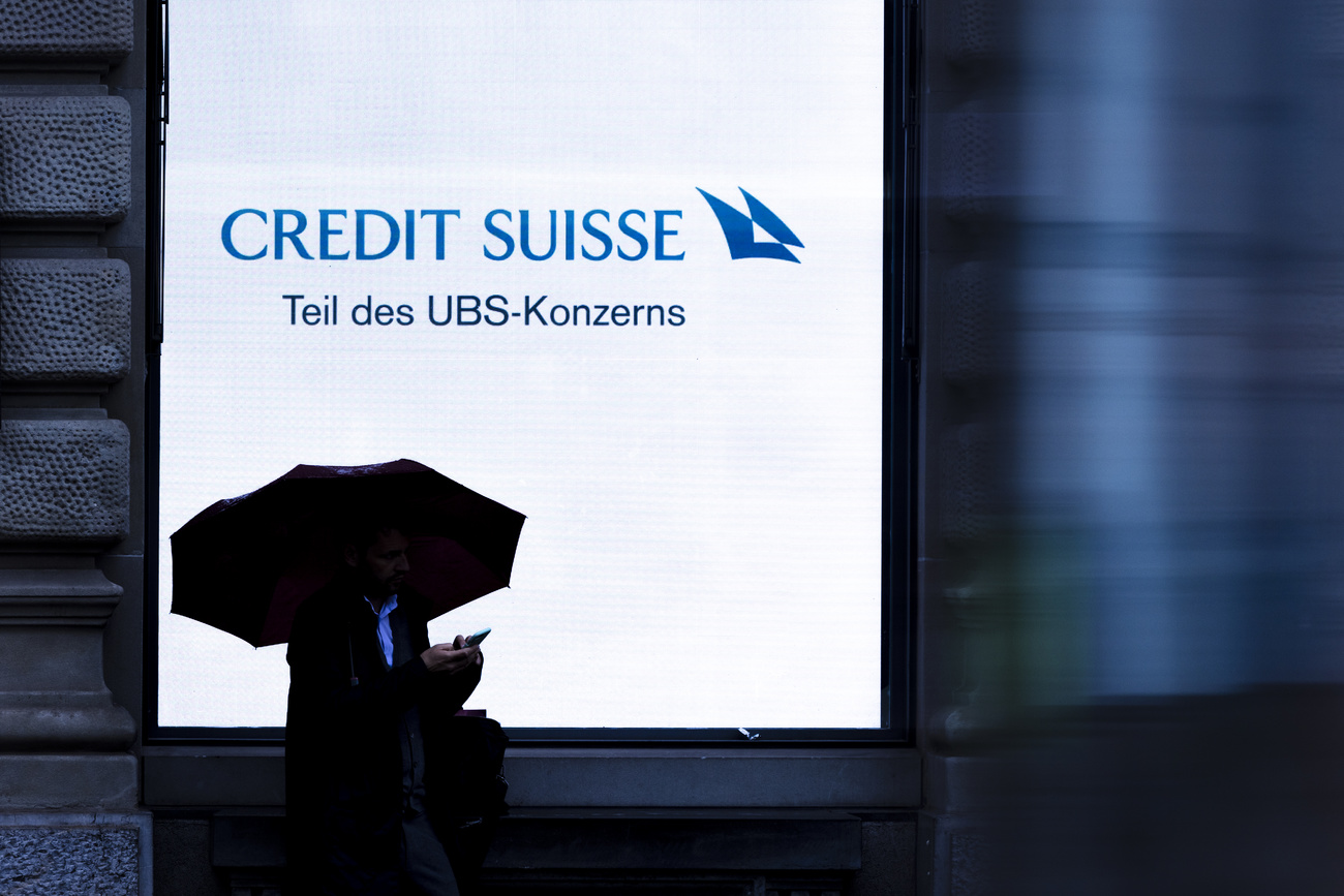 A person holding an umbrella is silhouetted against a screen of Swiss bank Credit Suisse with the words "Part of the UBS Group" at Zurich's Paradeplatz on Monday, June 3, 2024 in Zurich, Switzerland.