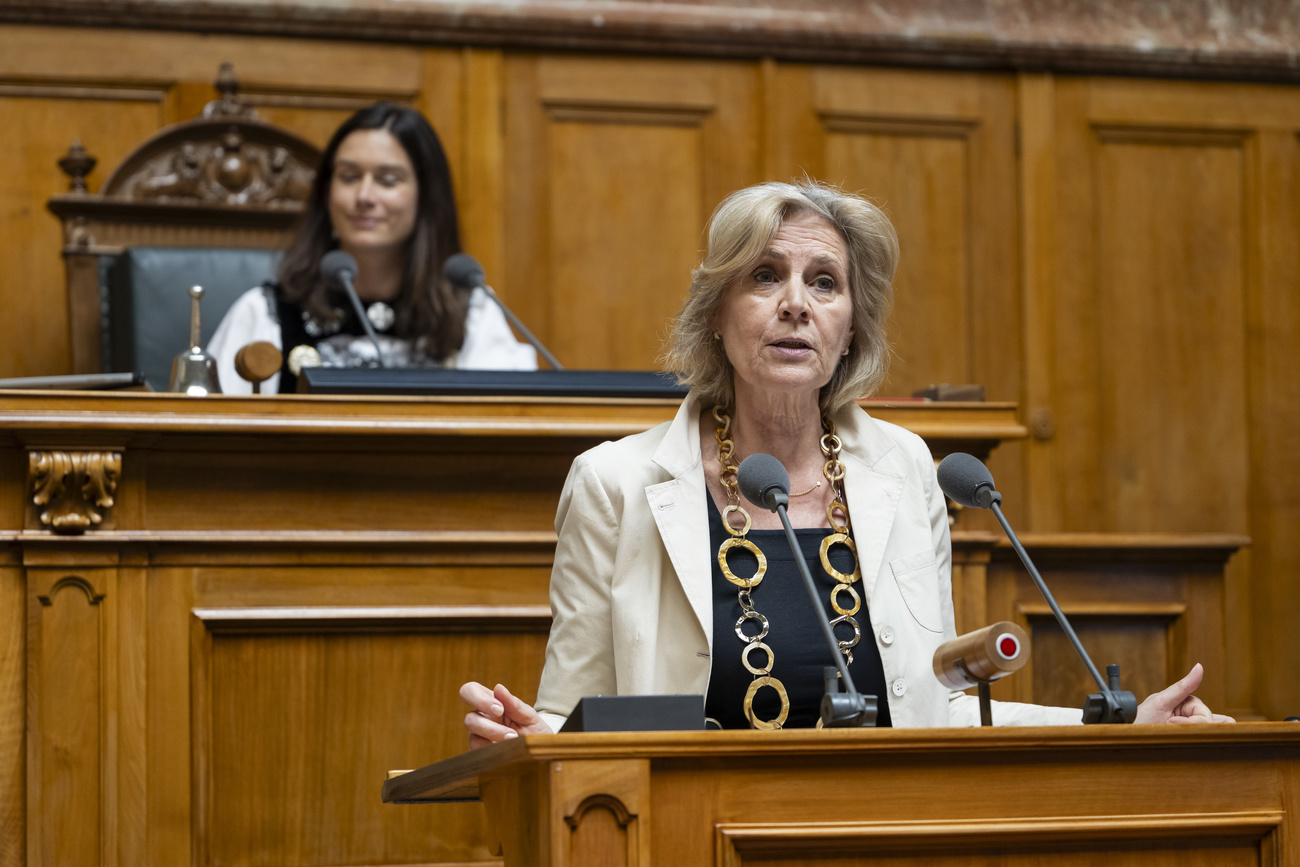 Patrizia von Falkenstein stands and speaks into a microphone at a wooden podium in Switzerland’s wood-panelled House of Representatives. She is wearing a black top, a cream jacket and a large necklace of gold rings interlocking. The outsides of her hands are resting, spread apart, on the podium. Vice-president Maja Riniker sits behind her.
