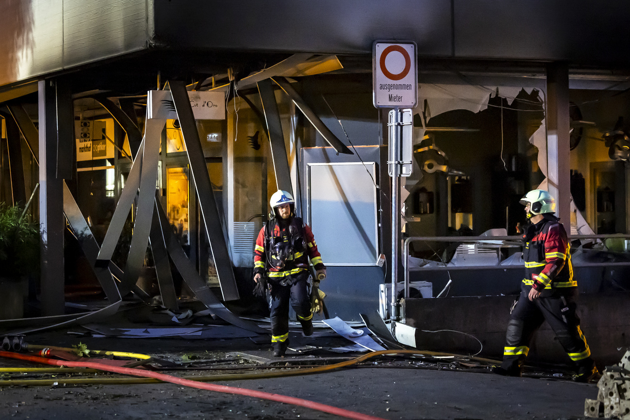 Two firefighters in full gear are shown in front of a building with broken windows and debris.