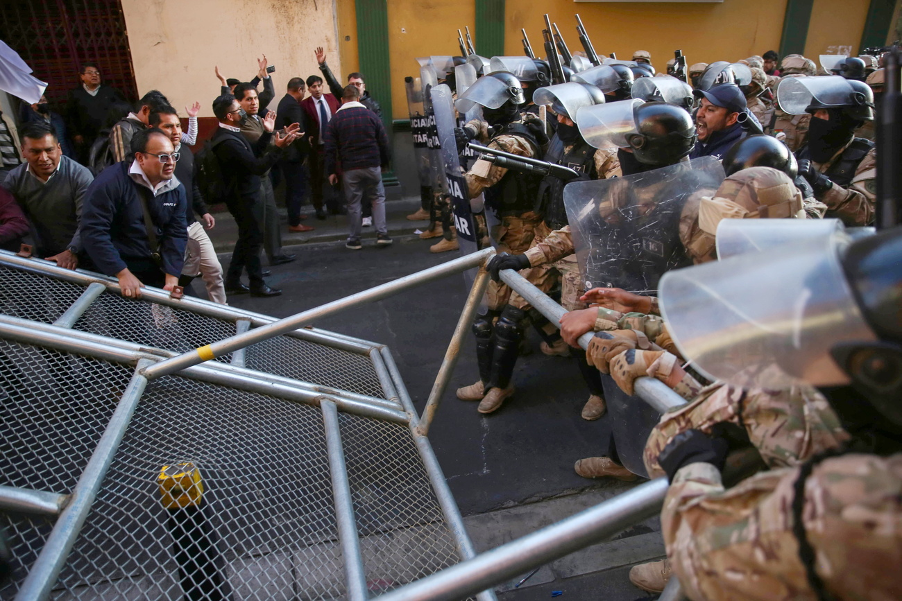 Soldiers in camouflage uniforms with black riot helmets and protective visors overturn a metal barrier.