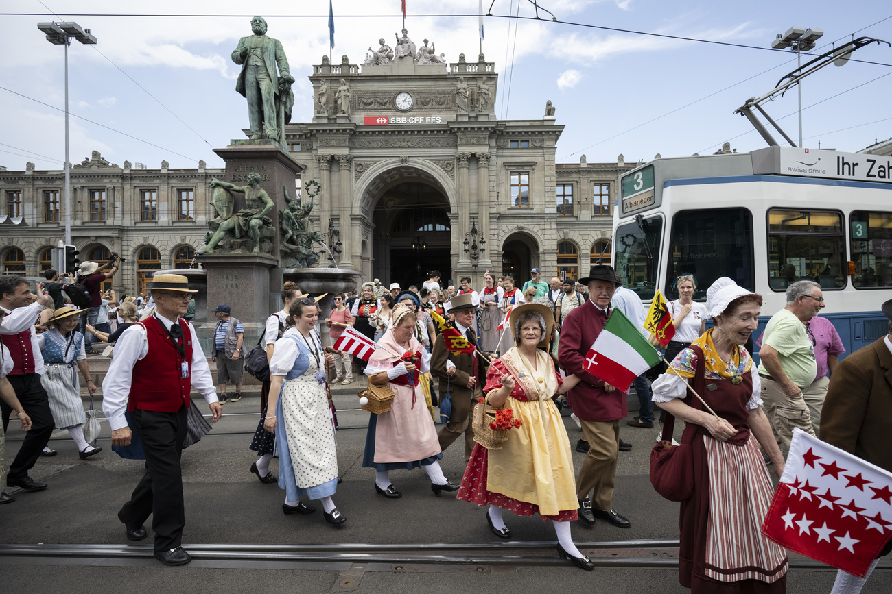 Women and men in traditional costume are shown walking on the street in front of Zurich's Hauptbahnhof.