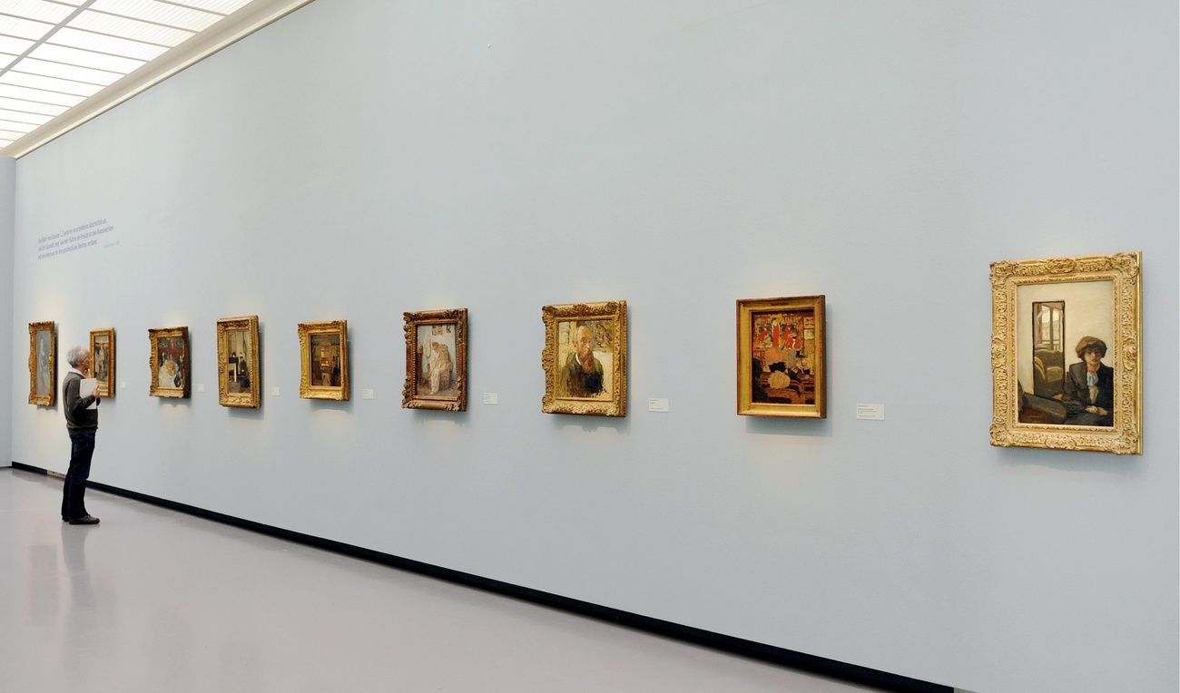 A visitor stands before a row of paintings on a wall at an art gallery.