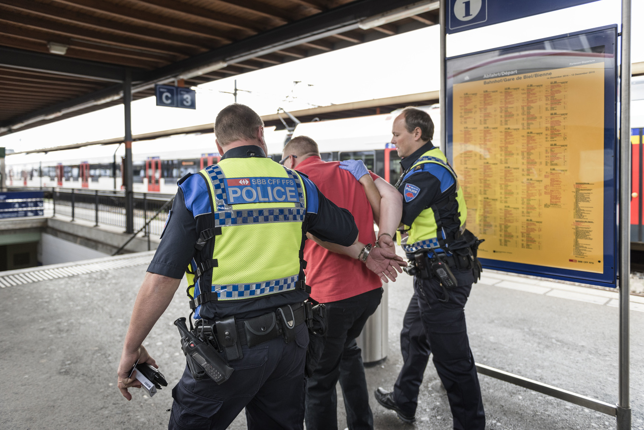 Two policemen in blue uniforms and yellow vests marked “POLICE” walk on either side of a man in a red shirt. The man between the two policemen is in handcuffs.