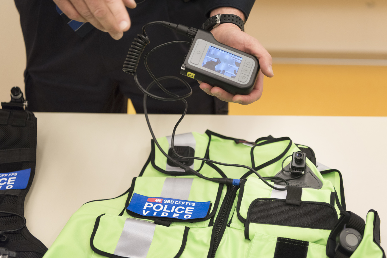 A yellow vest marked "police lays on a desk. hands are holding a palm sized rectangular body camera that is connect to a cable attached to the vest laying on the table.