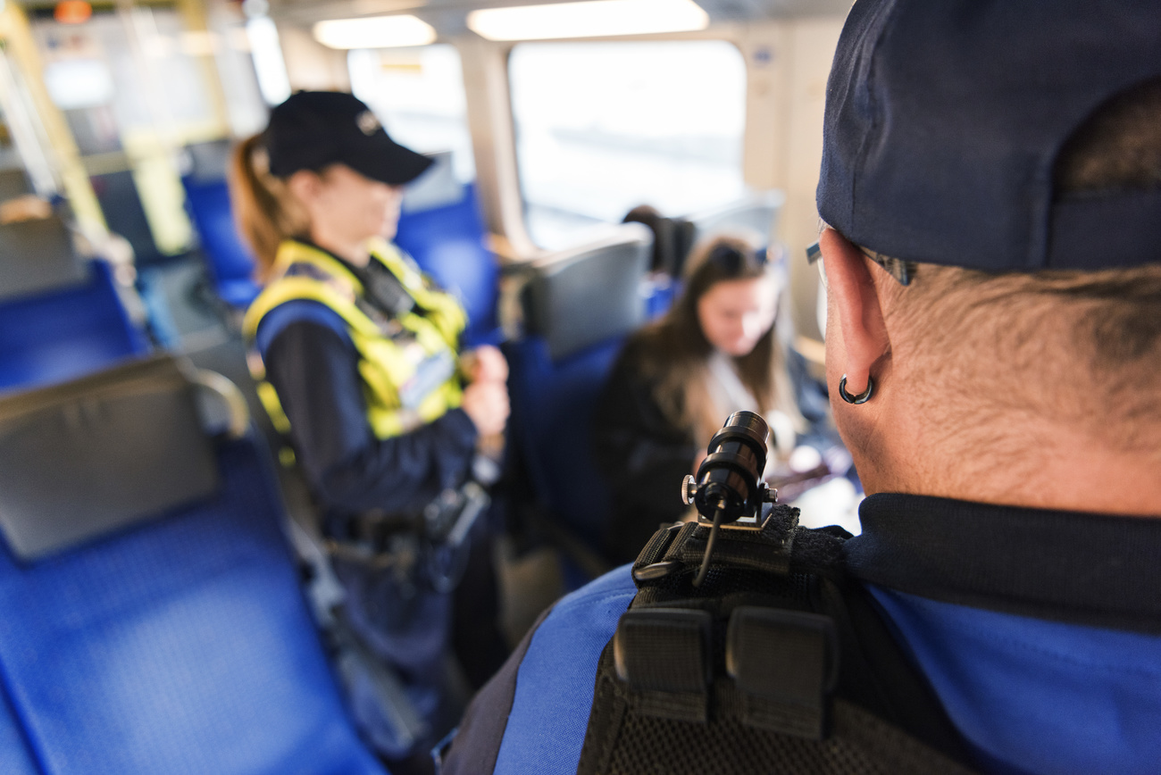 Two transport officers wearing black hats and yellow vests stop to talk to a woman sitting down. The seats of the train are blue and worn down in a seating marking.