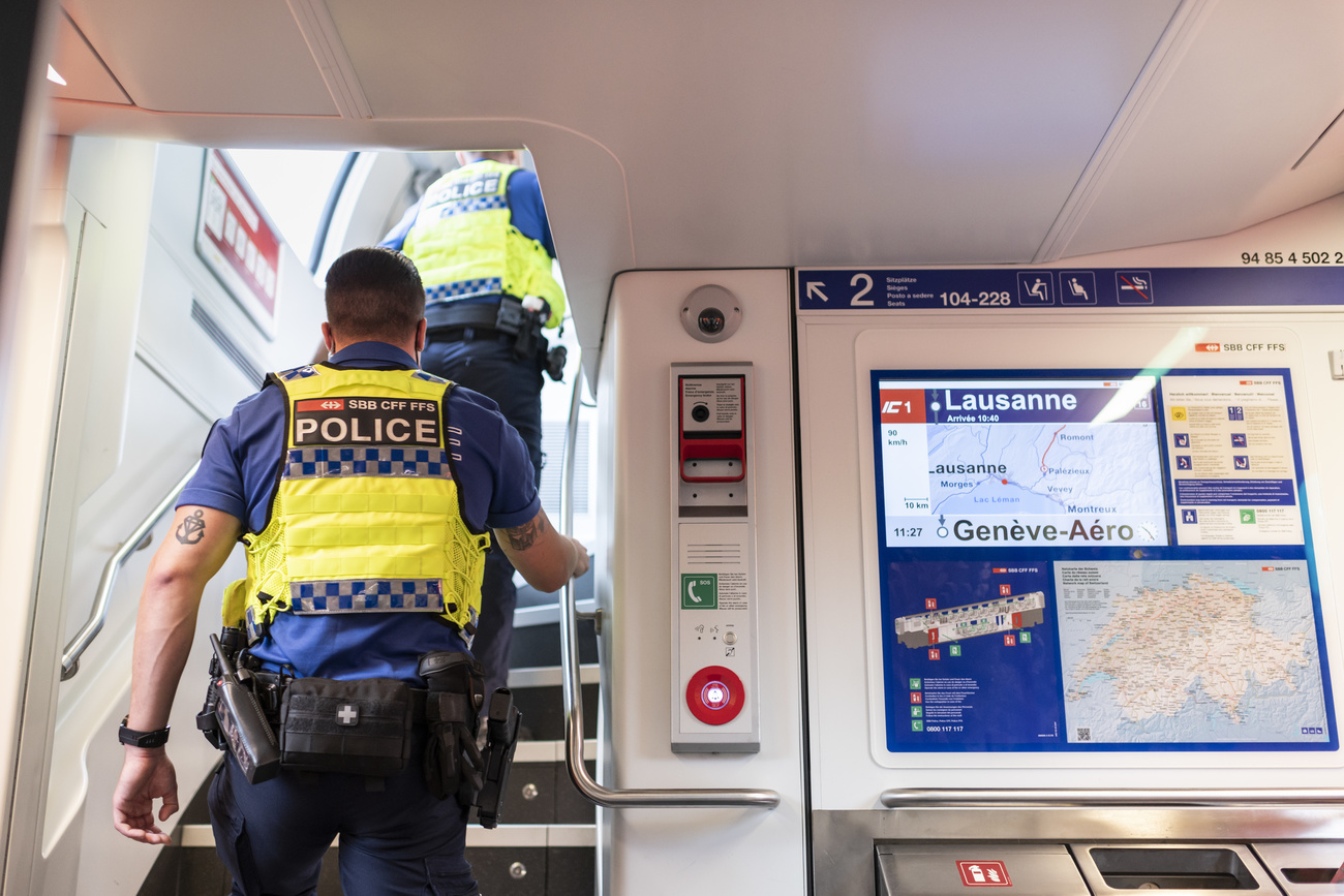 Two police men in blue uniforms and yellow vests marked “POLICE” walk up the stairs of a double decker train. A screen on the train says the train is going from Lausanne to Geneva Airport.