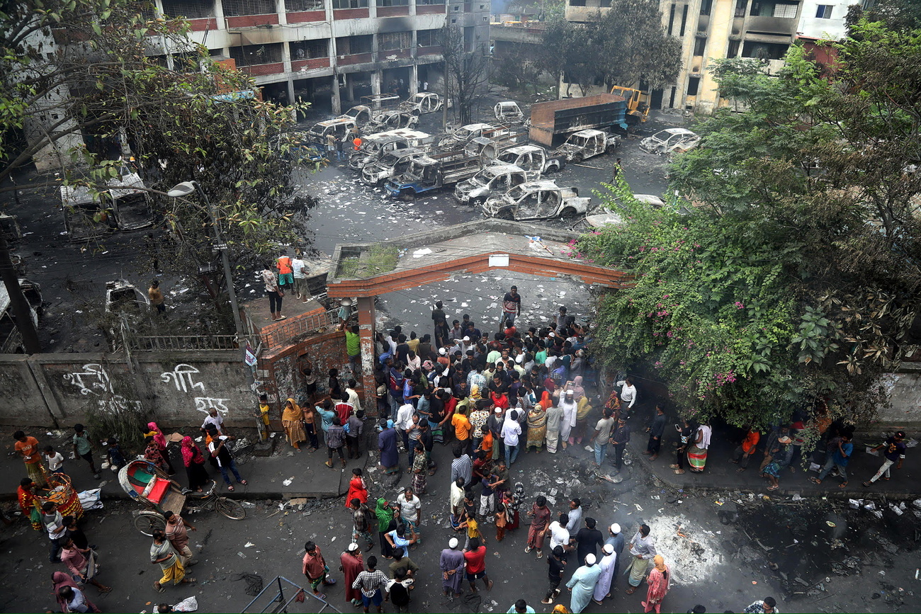 Multiple burned out cars are parked infront of a building that has signs of an explosion and fire. People stand at an entrance to the building.