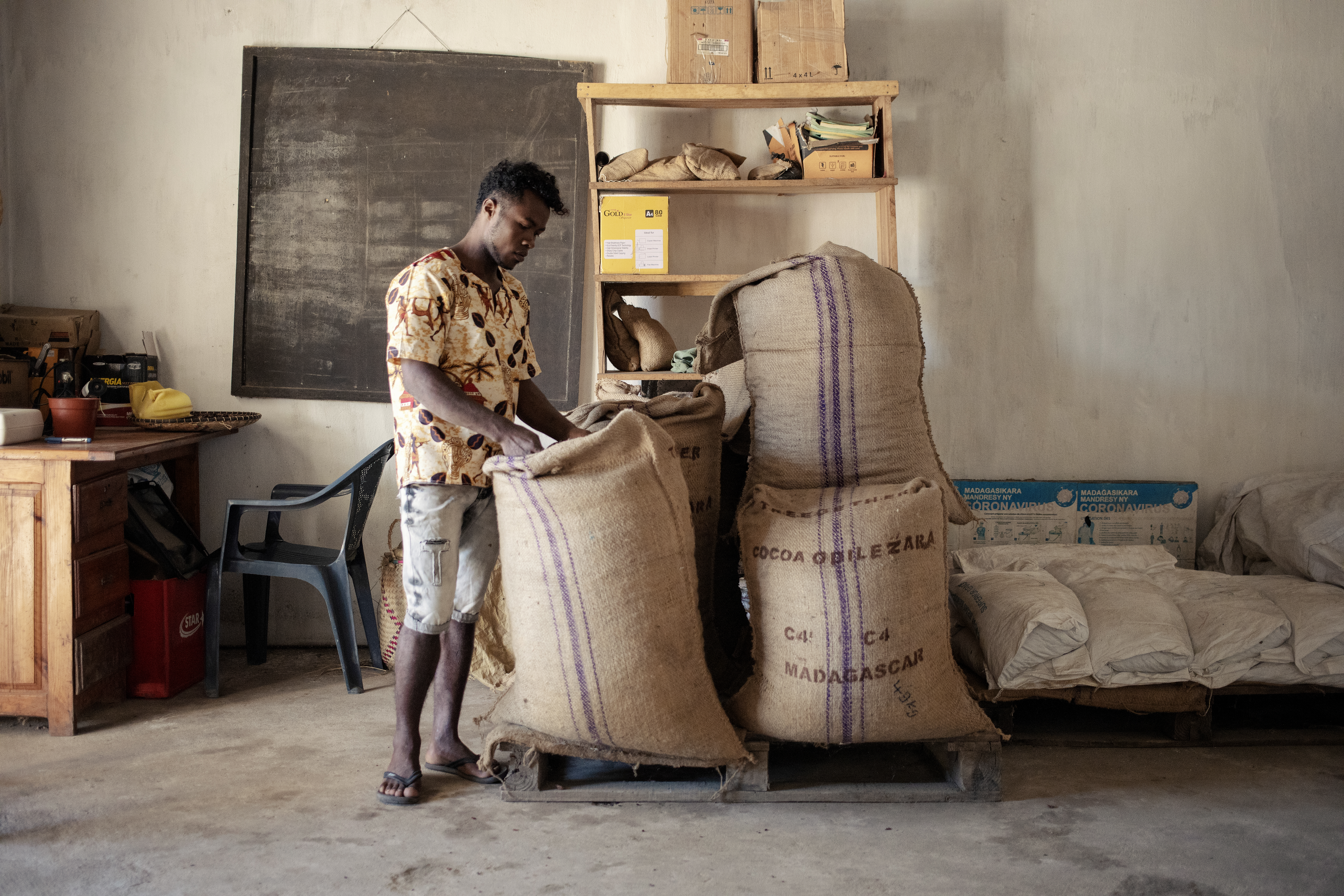 Person inspecting large burlap sacks labeled 'Cocoa Origin: Madagascar' in a modest storage room with shelves, bags, and scattered supplies.