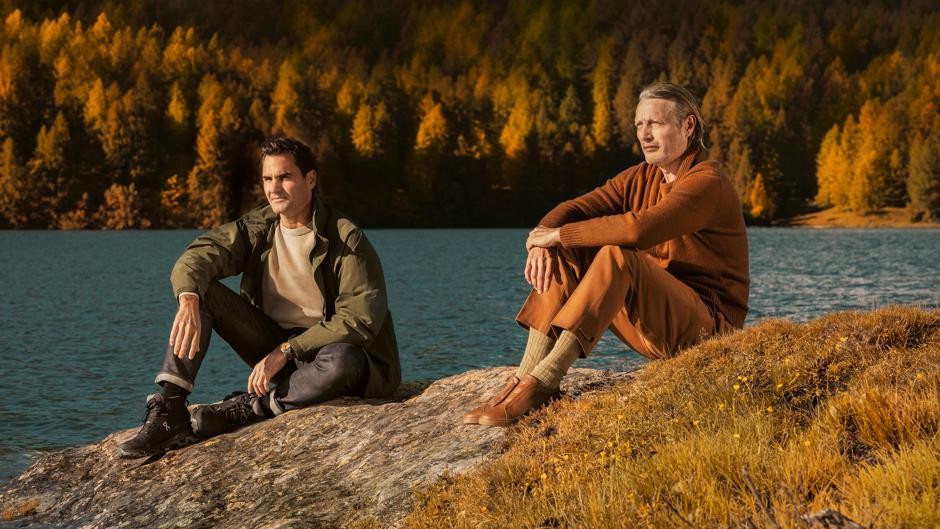 Two men sitting on a rock with a lake in the background. the trees are brown and orange displaying autumn