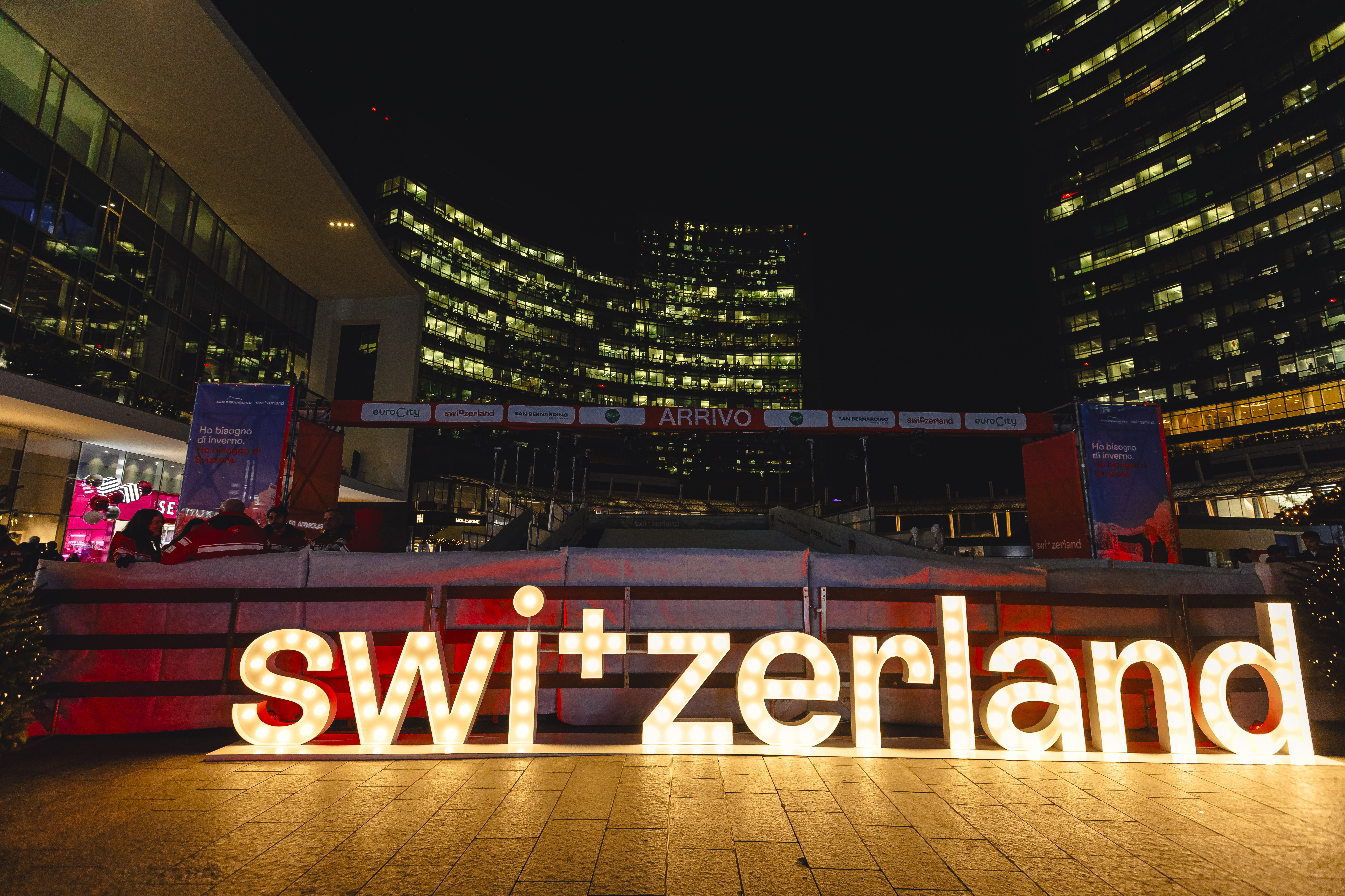 La scritta Switzerland illuminata con dietro i grattacilei di Piazza Gae Aulenti a Milano.