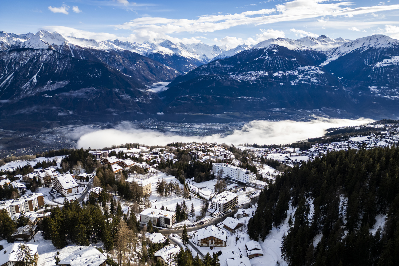 Aerial view of a snow-covered village in the Alps, surrounded by towering mountains and a valley filled with clouds.