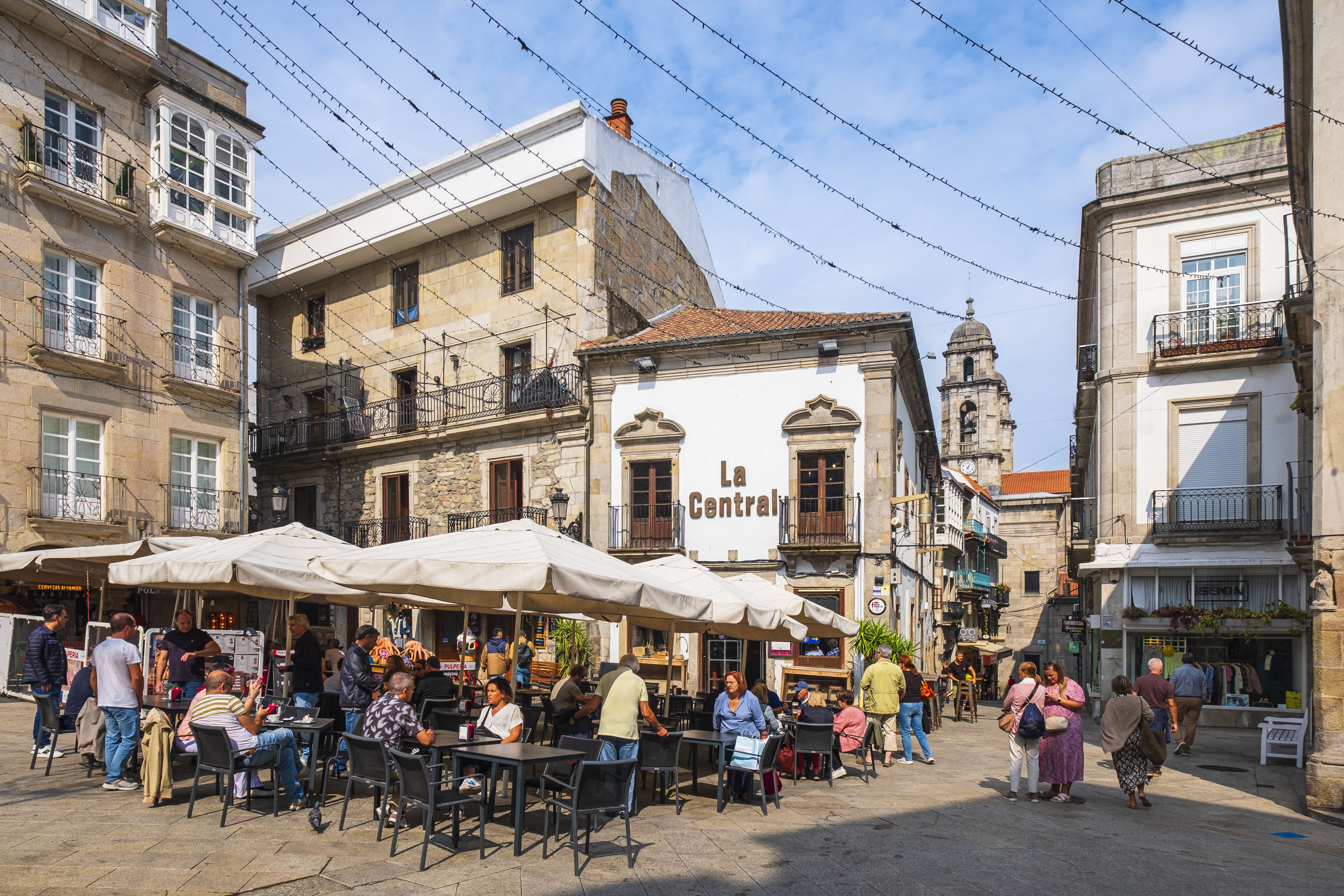 A bustling town square features outdoor cafes under umbrellas, with historic buildings and shoppers enjoying a sunny day.