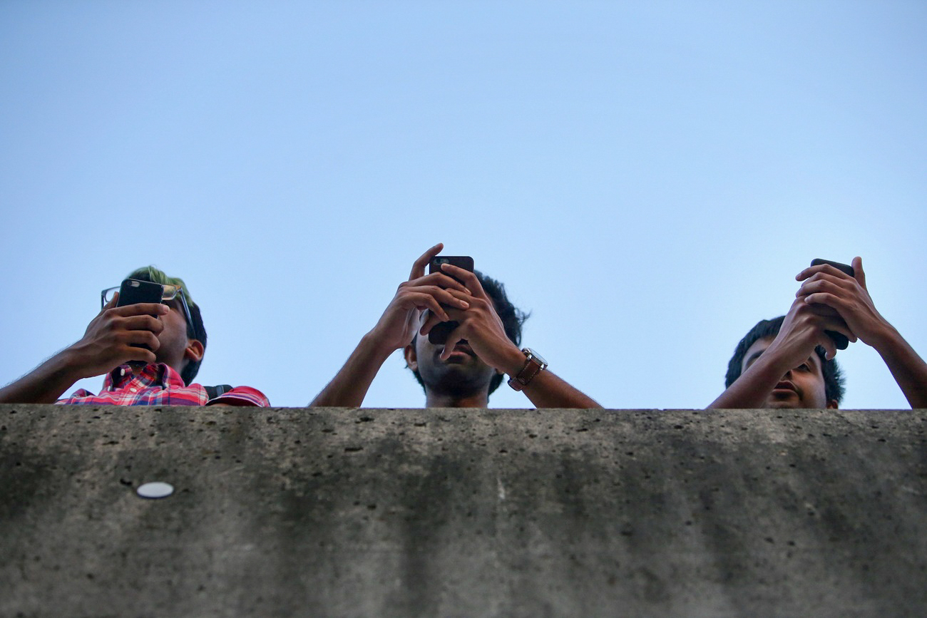 Three people, seen from below, hold smartphones while standing behind a concrete barrier against a clear blue sky.