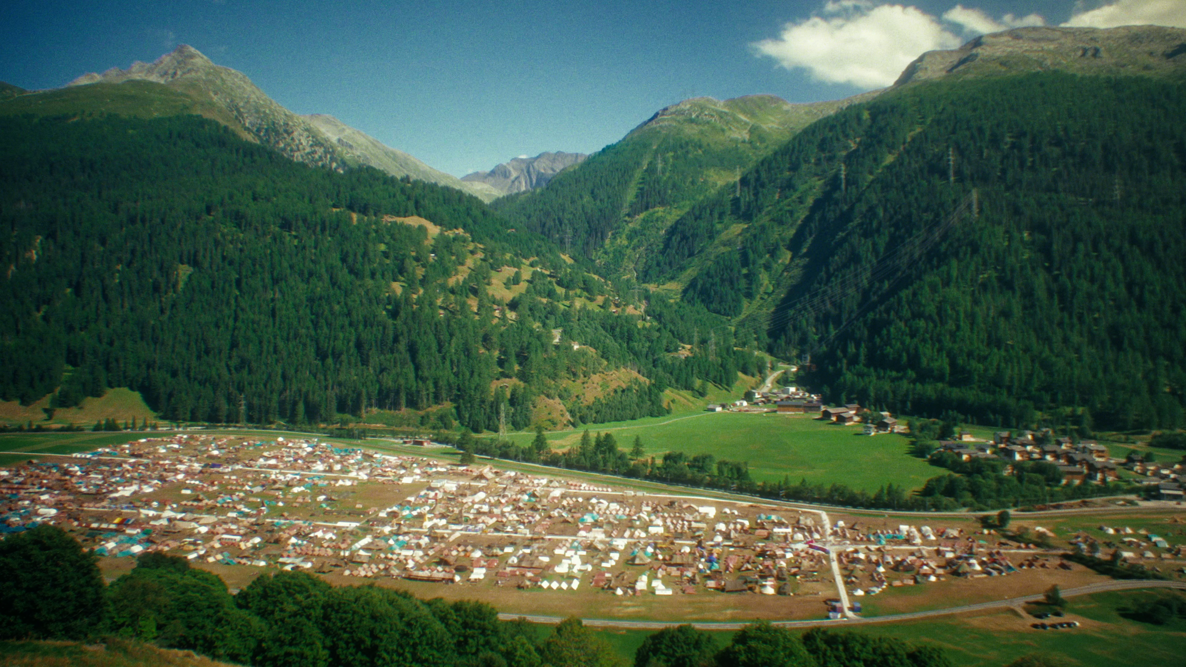 A panoramic view of a tent village nestled in a green valley, surrounded by mountains and lush forests.