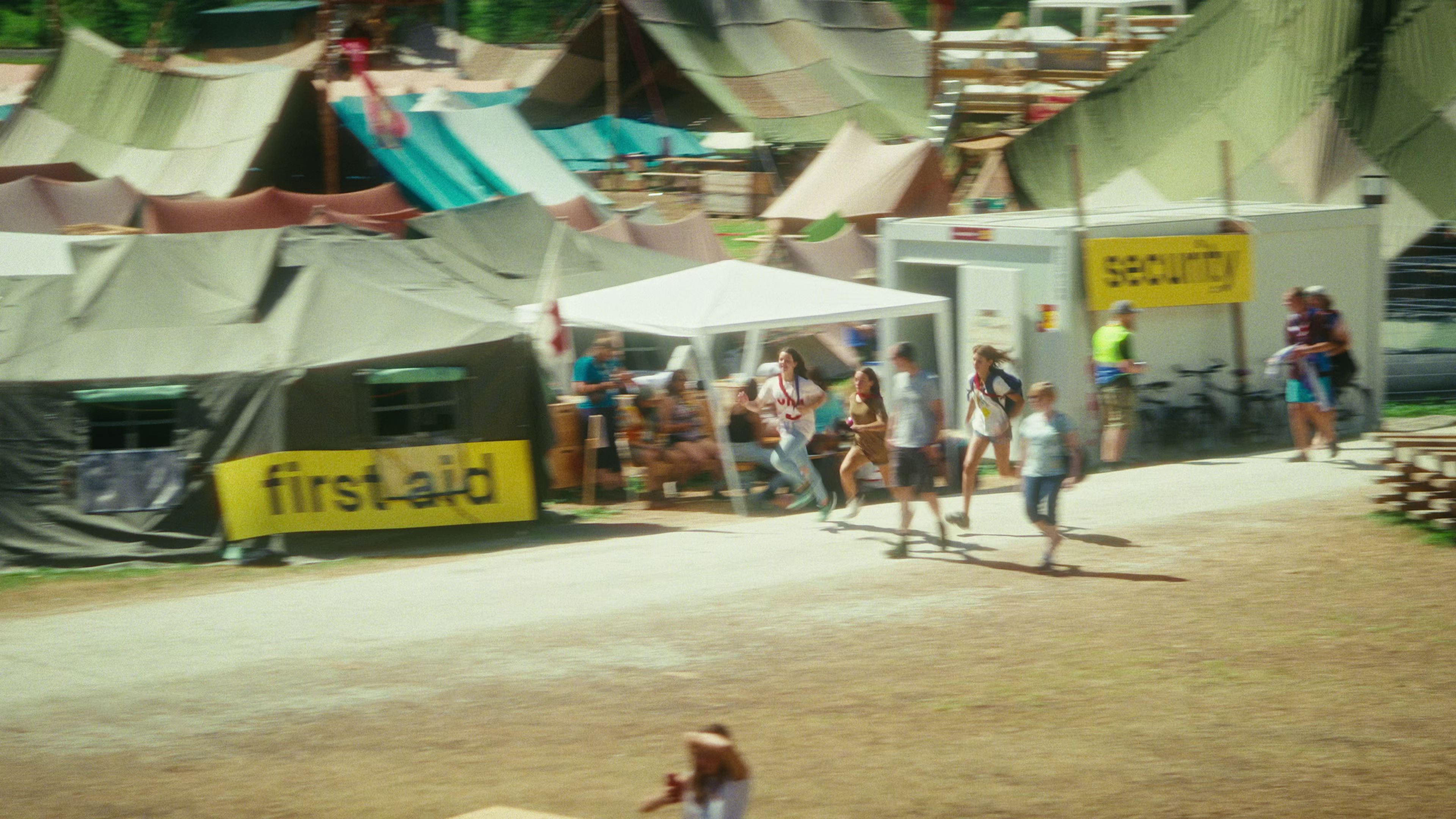 A bustling outdoor camp with various tents and a first aid station, as people walk and run along a dirt path.